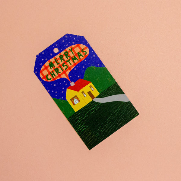 A Christmas-themed gift tag featuring a hand-painted style illustration of a yellow house, green hills, and a blue starry night sky with a red plaid banner reading 'MERRY CHRISTMAS'.