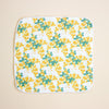 A square white cloth with rounded corners, featuring a repeating pattern of yellow flowers and green leaves, finished with a scalloped edge, laid flat on a light beige background.