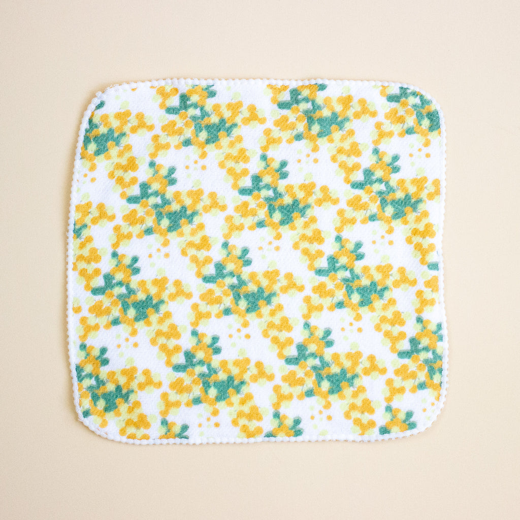 A square white cloth with rounded corners, featuring a repeating pattern of yellow flowers and green leaves, finished with a scalloped edge, laid flat on a light beige background.