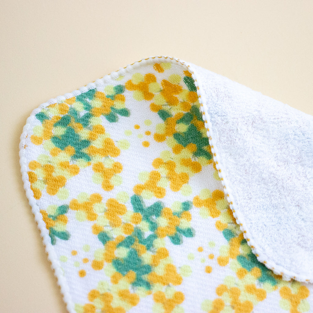 A close-up of a soft white cloth with a yellow and green floral pattern on one side and a plain white terry-like reverse side, laid on a light beige background.