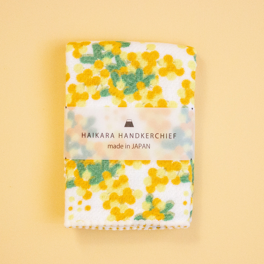 A folded white handkerchief with a yellow and green floral pattern, wrapped in a paper band that reads 'HAIKARA HANDKERCHIEF made in JAPAN', set against a solid pale yellow background.