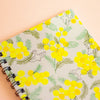 A spiral-bound notebook with a light grey-coloured cover decorated with a repeating pattern of bright yellow mimosa flowers, green leaves, and simple bird illustrations, shown on a solid peach background.