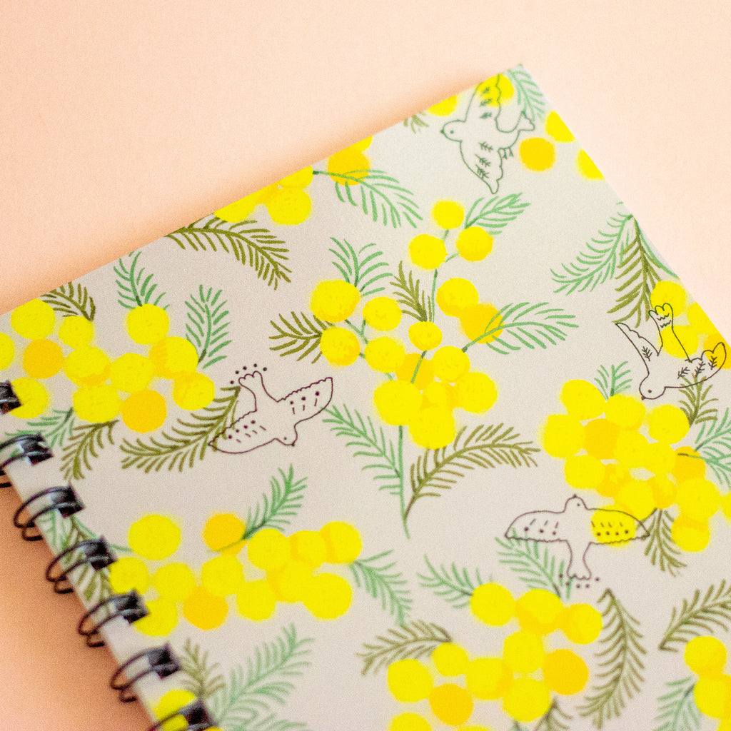 A spiral-bound notebook with a light grey-coloured cover decorated with a repeating pattern of bright yellow mimosa flowers, green leaves, and simple bird illustrations, shown on a solid peach background.