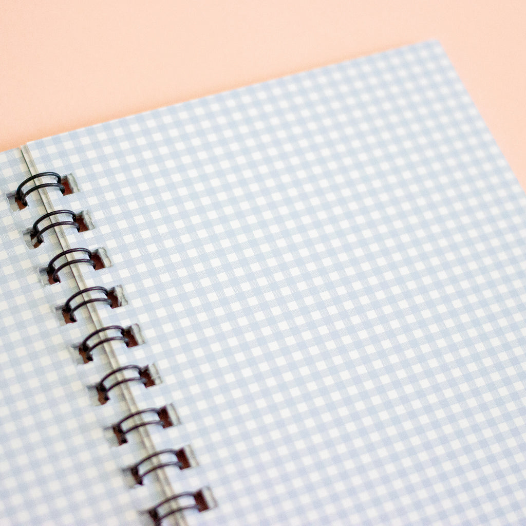 A close-up of a spiral-bound notebook with a light blue and white gingham patterned cover, bound with black metal rings on a peach-coloured background.