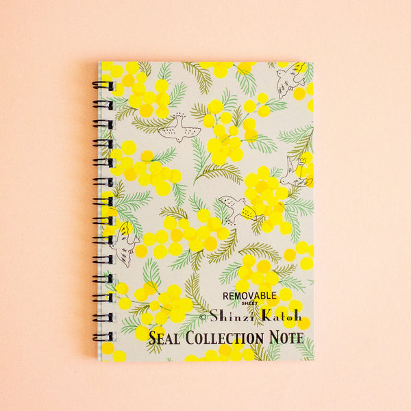 A spiral-bound notebook titled 'Shinzi Katoh Seal Collection Note' featuring a yellow and green floral pattern on a light background, set against a solid peach-pink backdrop.