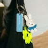 A close-up of a black shoulder bag with a keychain attached. The keychain features a white rabbit character reading a blue book, a bright neon green flower charm, and a blue plastic ruler tag.