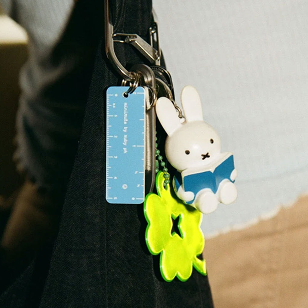A close-up of a black shoulder bag with a keychain attached. The keychain features a white rabbit character reading a blue book, a bright neon green flower charm, and a blue plastic ruler tag.