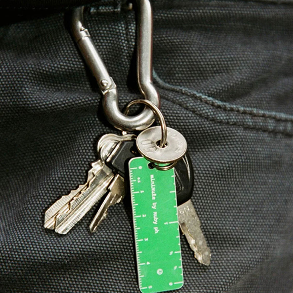 A close-up of a silver carabiner holding several keys and a bright green ruler keychain with white measurement markings, attached to a black pants belt loop.