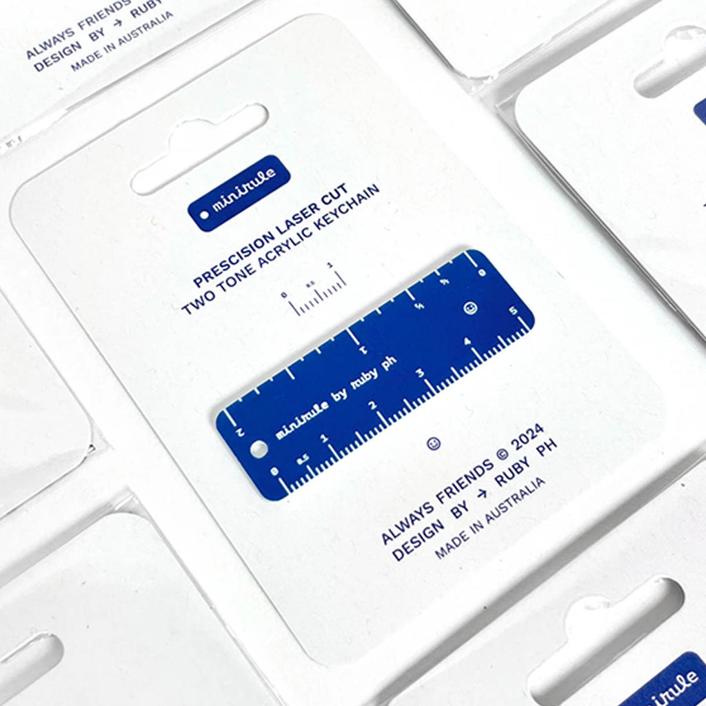 A close-up of a blue two-tone acrylic keychain ruler inside its white retail packaging, with printed text including 'PRECISION LASER CUT TWO TONE ACRYLIC KEYCHAIN' and 'ALWAYS FRIENDS by Ruby PH'.