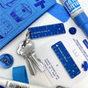 A close-up flat lay of blue matching stationery accessories on a white surface. The set includes a small ruler, a keychain with two keys, alongside blue pens and a blue notebook.