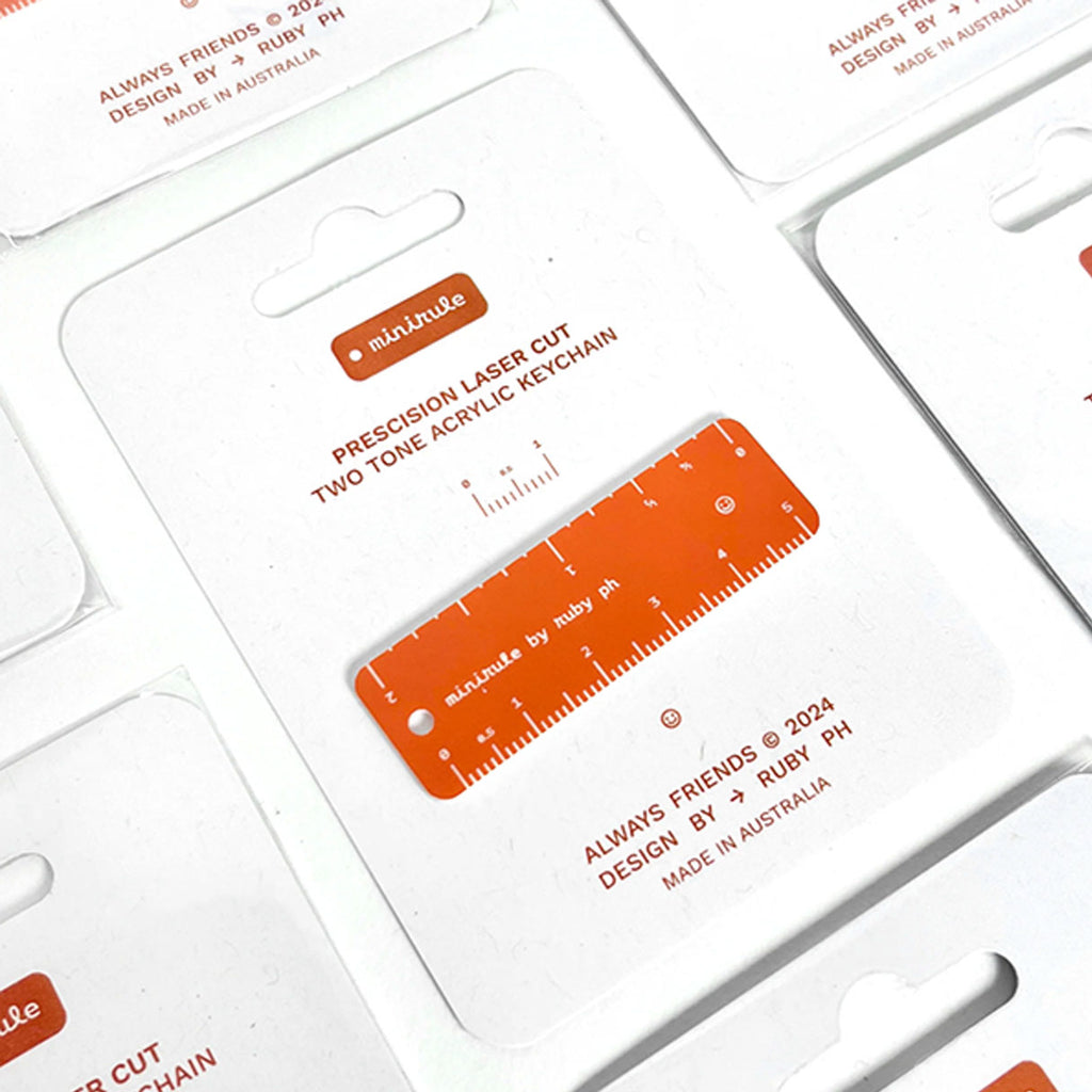 A close-up of several white acrylic keychain cards arranged in a grid, each featuring an orange laser-cut ruler keychain with printed measurement markings and small text including 'PRECISION LASER CUT TWO TONE ACRYLIC KEYCHAIN'.