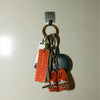 A set of keys with a silver metal ring and hook, featuring a small orange ruler, and miniature orange clogs, hanging on a plain light beige wall.