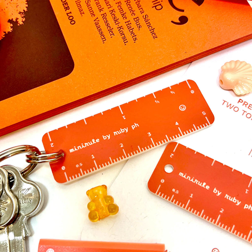 Orange plastic pocket rulers with white measurement markings and the text 'minirule by ruby ph' printed on them, shown attached to a keyring with keys on a white surface alongside small decorative items.