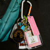 A close-up of a carabiner holding a keychain set with a round black metal medallion, a pink ruler, a clear ID window card, and other small keys, shown attached to a teal bag against a dark fabric background.