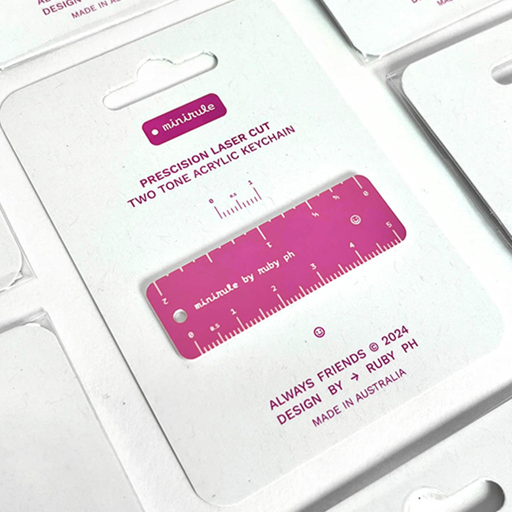A precision laser-cut two-tone acrylic keychain ruler, shown in its original white packaging. The keychain features a vibrant magenta pink and white colour combination with measurement markings.