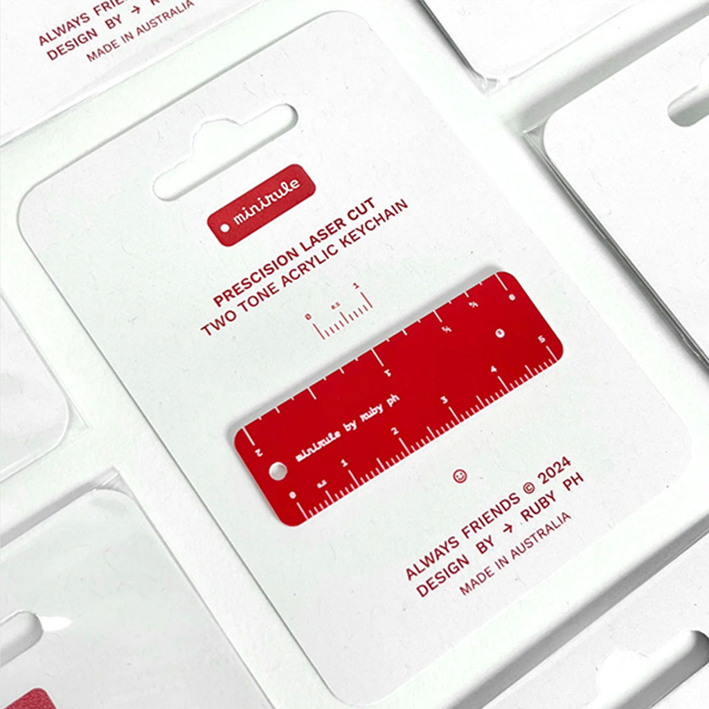 A red acrylic precision laser-cut ruler keychain displayed on its white backing card, which features product text and branding.