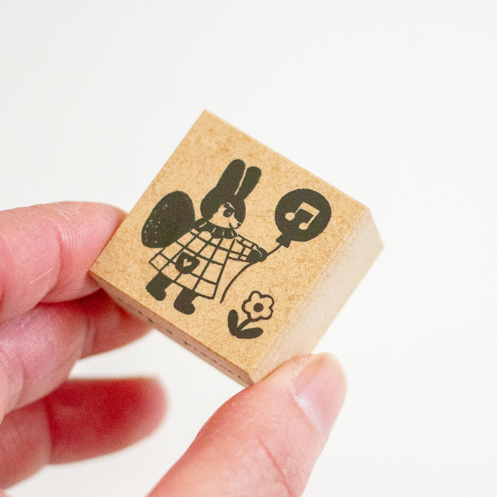 A hand holding a small square wooden stamp block featuring a black printed illustration of a rabbit holding a music note balloon.