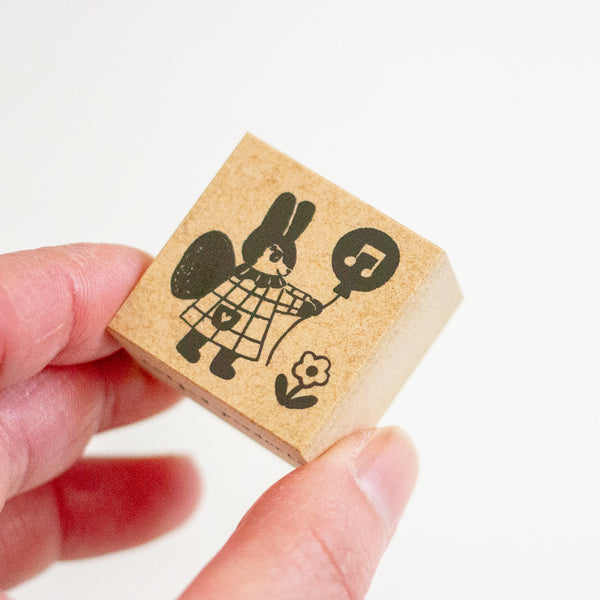 A hand holding a small square wooden stamp block featuring a black printed illustration of a rabbit holding a music note balloon.