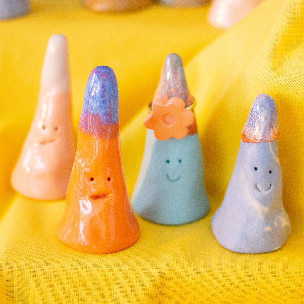 A closeup picture of 4 ring holders, with smiling faces, different colours and glazes, and one is holding a flower ring. 