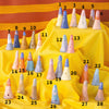 A photo of the entire collection of ceramic ring holders. In this photo, each ring holder is numbered for ease of choosing. Ring holders are placed on a yellow tiered fabric stand, with a striped background. Ring holders are cylindrical in shape, multiple colours, and feature different faces and glazes. 