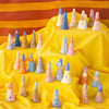 A photo of the entire collection of ceramic ring holders. Ring holders are placed on a yellow tiered fabric stand, with a striped background. Ring holders are cylindrical in shape, multiple colours, and feature different faces and glazes. 