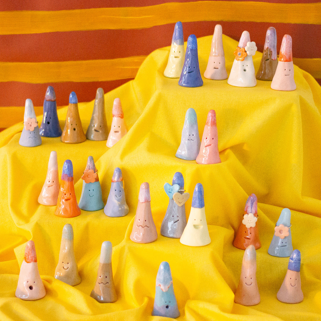 A photo of the entire collection of ceramic ring holders. Ring holders are placed on a yellow tiered fabric stand, with a striped background. Ring holders are cylindrical in shape, multiple colours, and feature different faces and glazes. 