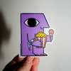 A hand holds a die-cut sticker featuring a purple cartoon character with a single eye holding popcorn and a 3D glasses, against a plain light background.