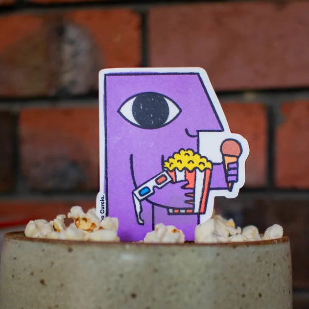 A ceramic popcorn bowl with a speckled beige exterior and a dark blue base, topped with popcorn and a purple cartoon pick with an eye and popcorn cup, set on a wooden table against a brick wall background.