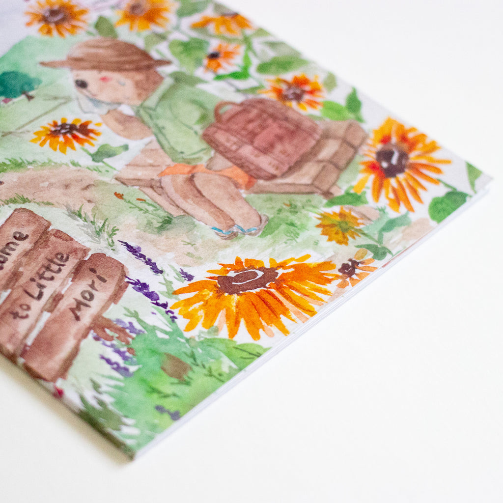 A square watercolour-style book featuring a whimsical illustration of an otter sitting on the bench with a backpack, surrounded by sunflowers and green leaves, photographed on a clean white background.