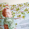 An open illustrated book showing a watercolour-style scene with a brown otter wearing a green shirt among leafy branches, with printed text on the right page.
