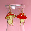 A pair of dangling mushroom-shaped earrings with red caps, white spots, and cream bodies, hanging from gold hooks in front of a pink glass container.