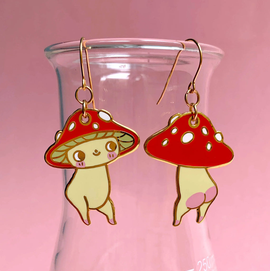 A pair of dangling mushroom-shaped earrings with red caps, white spots, and cream bodies, hanging from gold hooks in front of a pink glass container.