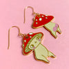 A pair of mushroom-shaped dangling earrings with cute cartoon faces, featuring red caps with white spots, pale yellow-green bodies, and gold-tone hooks, displayed on a solid pink background.