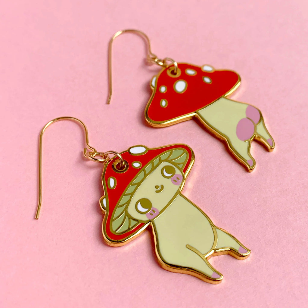 A pair of mushroom-shaped dangling earrings with cute cartoon faces, featuring red caps with white spots, pale yellow-green bodies, and gold-tone hooks, displayed on a solid pink background.