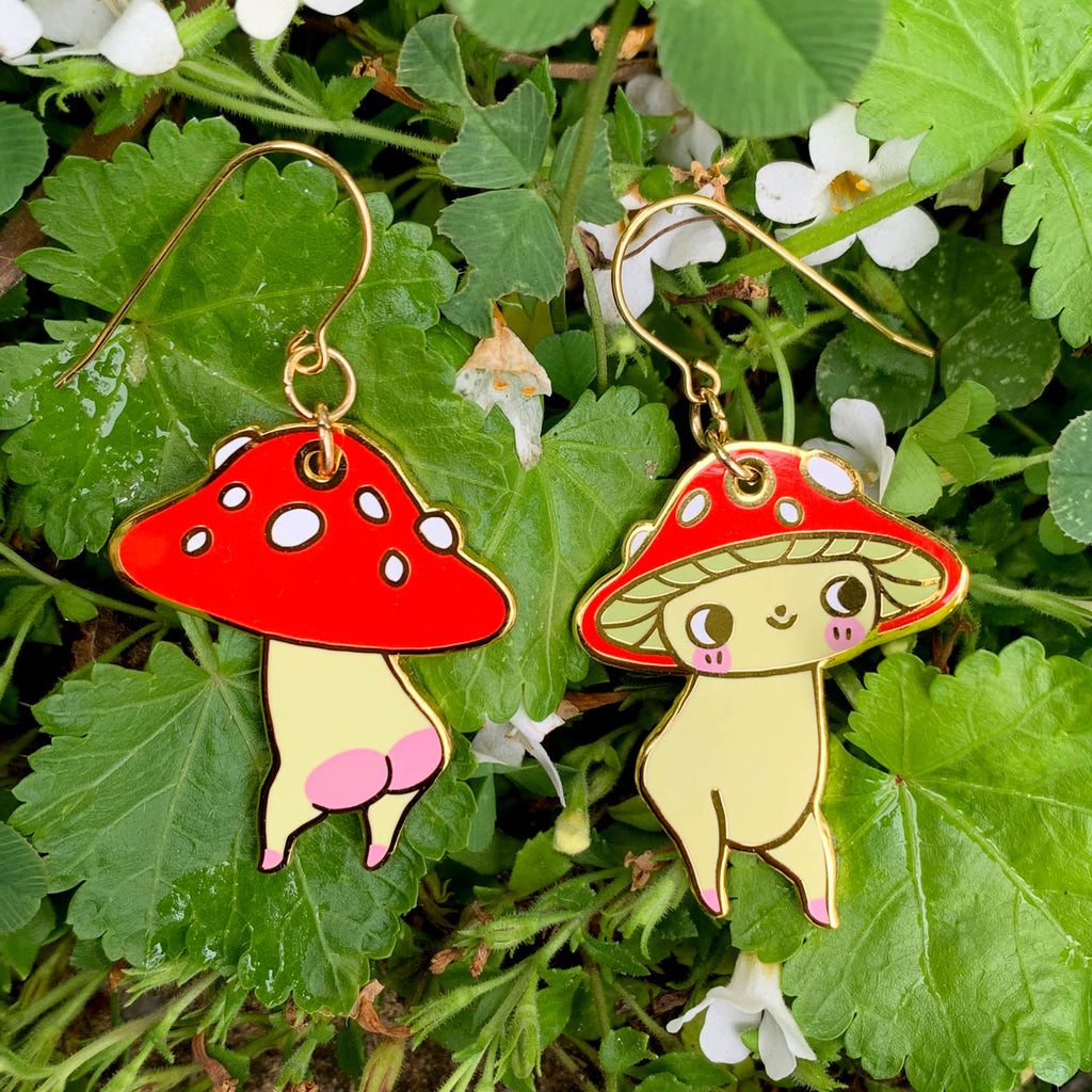 A pair of dangling mushroom-shaped earrings hanging outdoors among green leaves and small white flowers. The earrings have red caps with white spots, pale yellow bodies, and gold-tone hooks.