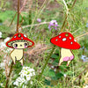 Two cartoon mushroom-shaped earrings hanging from a plant stem in an outdoor garden setting with green stems and small white and purple flowers.