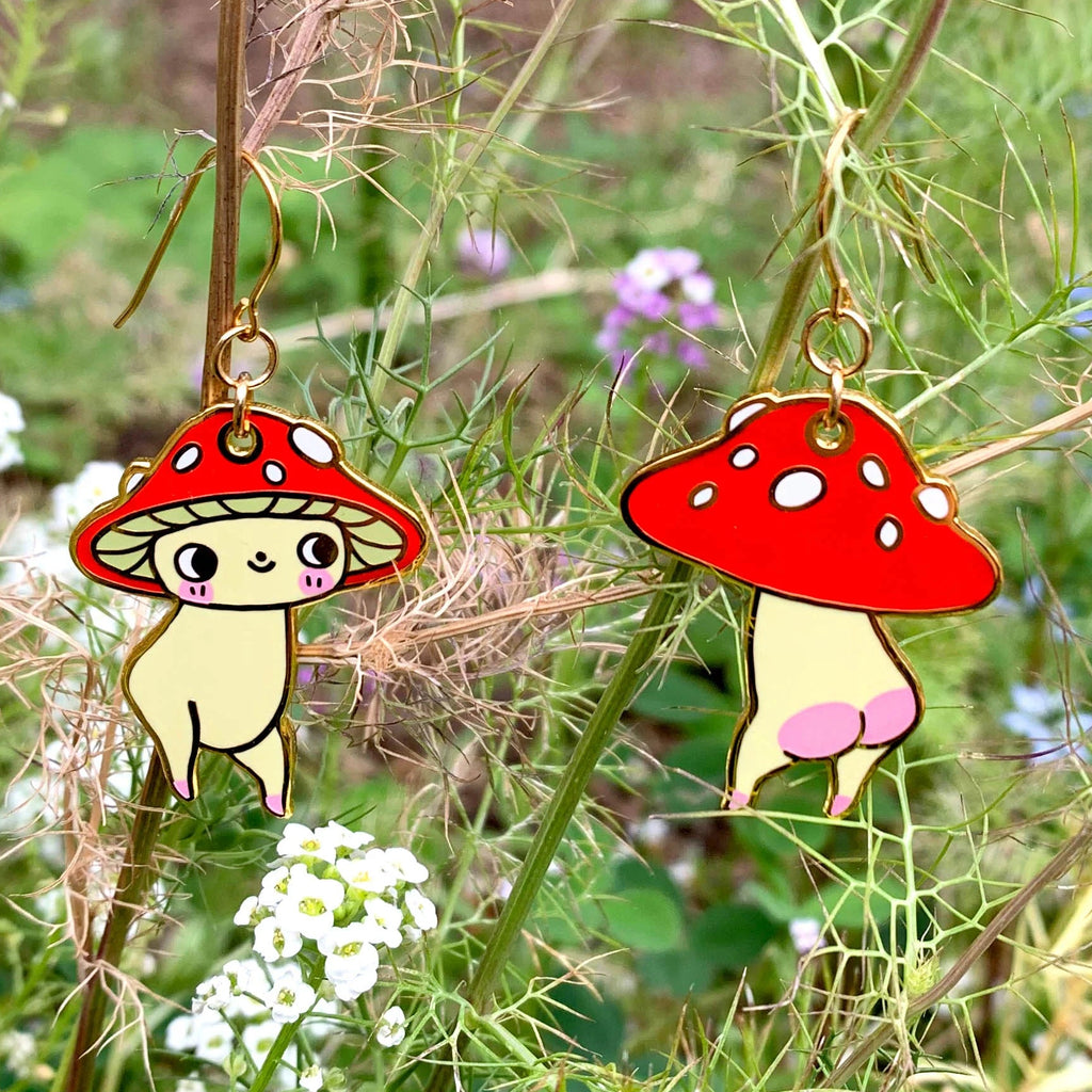 Two cartoon mushroom-shaped earrings hanging from a plant stem in an outdoor garden setting with green stems and small white and purple flowers.