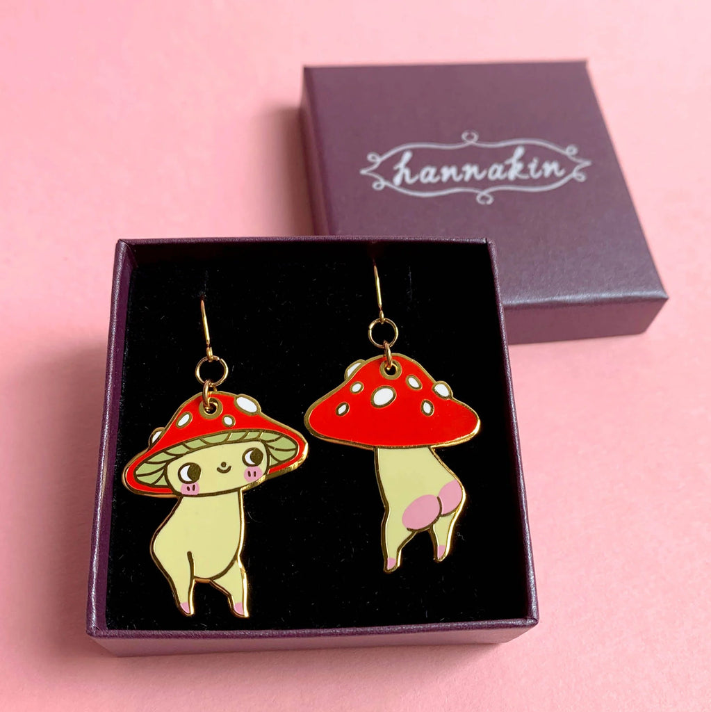 A pair of dangling mushroom-shaped earrings displayed in an open black jewelry box, with the brand name hannakin visible on the purple lid behind it. The earrings feature red-and-white spotted mushroom caps and pale yellow bodies with pink accents, set against a soft pink background.