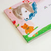 A close-up of a notepad, featuring a whimsical illustration of a dog's face, a small orange teddy bear, flowers, and a green grassy corner on a light pink background.