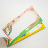 A pastel notepad with whimsical animal illustrations, featuring a white dog on a green field with a pink floral border, photographed on a clean white background.