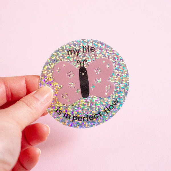 A hand holding a round glitter sticker featuring a pink butterfly and the text 'my life is in perfect flow' against a solid pink background.
