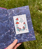 A hand holding an open book, inside there is a small paper stuck on it with illustrations of mushrooms with words 'This book belongs to'.