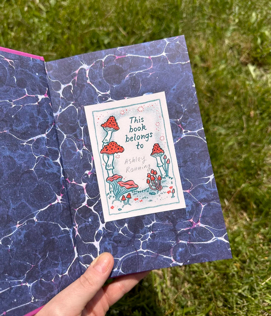 A hand holding an open book, inside there is a small paper stuck on it with illustrations of mushrooms with words 'This book belongs to'.
