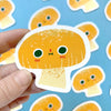 Hand holding a cute orange character magnet with a blue background featuring more of these magnets.