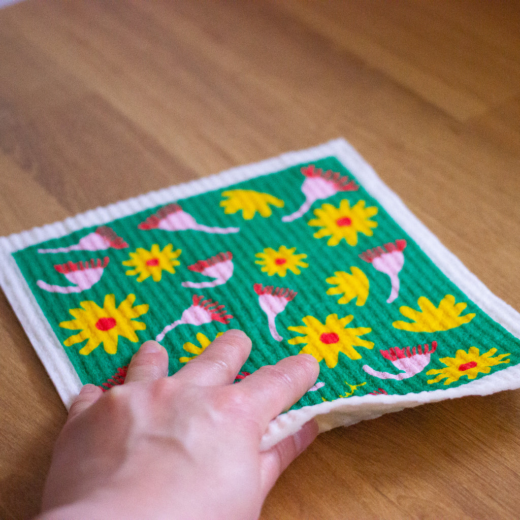 A picture of a hand using the shuh and spruce native flora dishcloth to wipe a bench. 