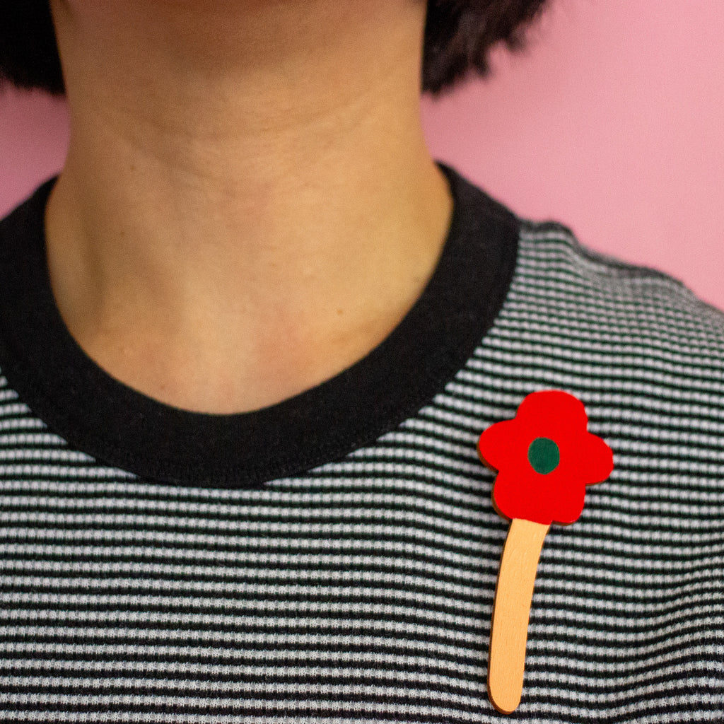 Person wearing a black and white striped shirt with a red flower on a stick attached to their collarbone against a pink background.