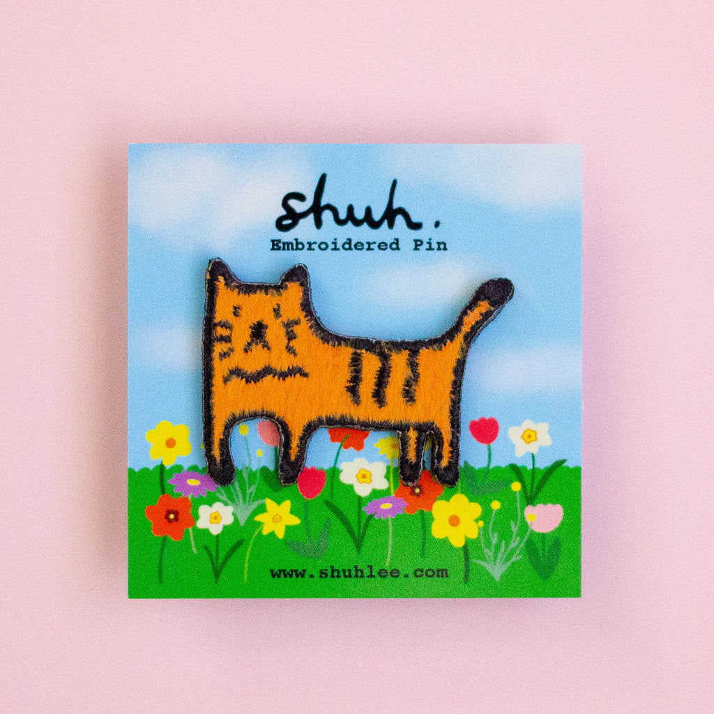 Embroidered pin of a cat in a field with 'shuh' branding on a pink background