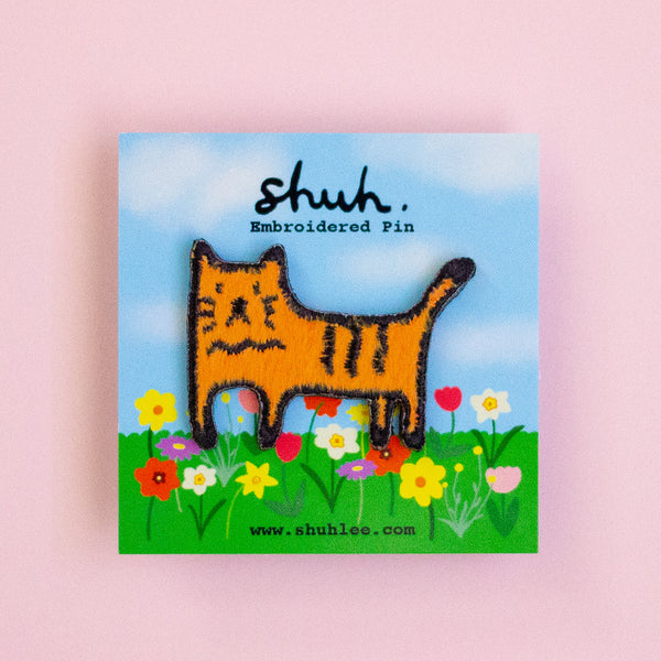 Embroidered pin of a cat in a field with 'shuh' branding on a pink background