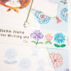 A close-up of a writing set by Aiko Fukuwa, featuring a decorative paper strip with colourful hand-drawn flowers and Japanese text on a white background.