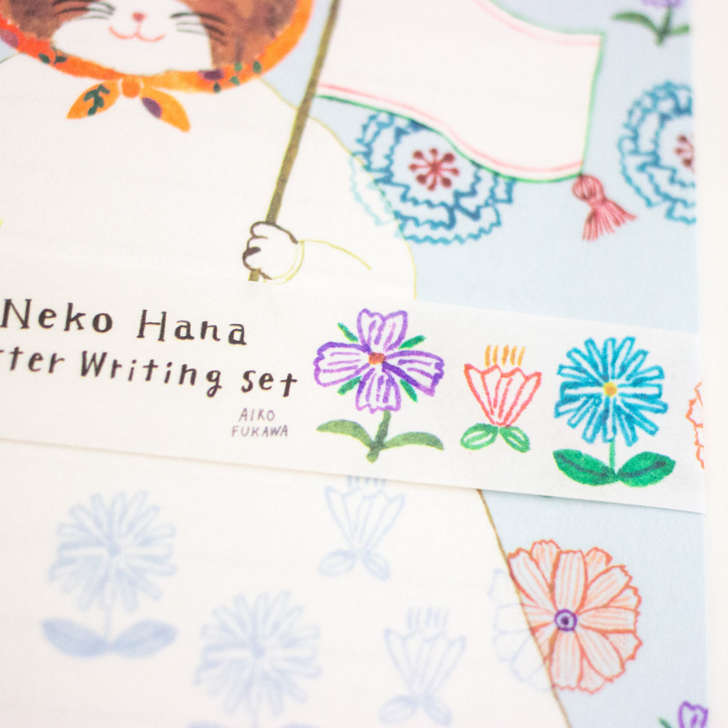 A close-up of a writing set by Aiko Fukuwa, featuring a decorative paper strip with colourful hand-drawn flowers and Japanese text on a white background.